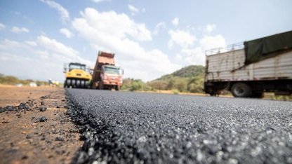 A road construction site showing a freshly paved asphalt surface with heavy machinery including a steam roller and a dump truck in the background under a blue sky.