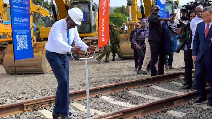 A wide shot of an active construction site for a railway station with heavy machinery and steel reinforcements visible against a clear sky.