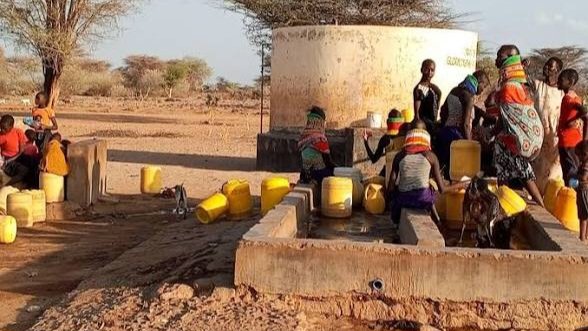 Local residents in an arid region queue with yellow jerrycans around a large concrete water storage tank and a dry borehole point.