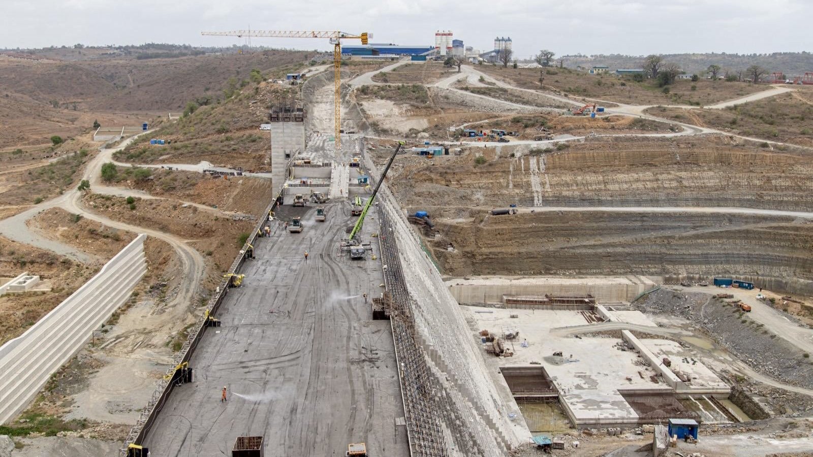 A wide-angle view of the Mwache Dam construction site showing heavy machinery compacting layers of roller-compacted concrete on the main dam wall.