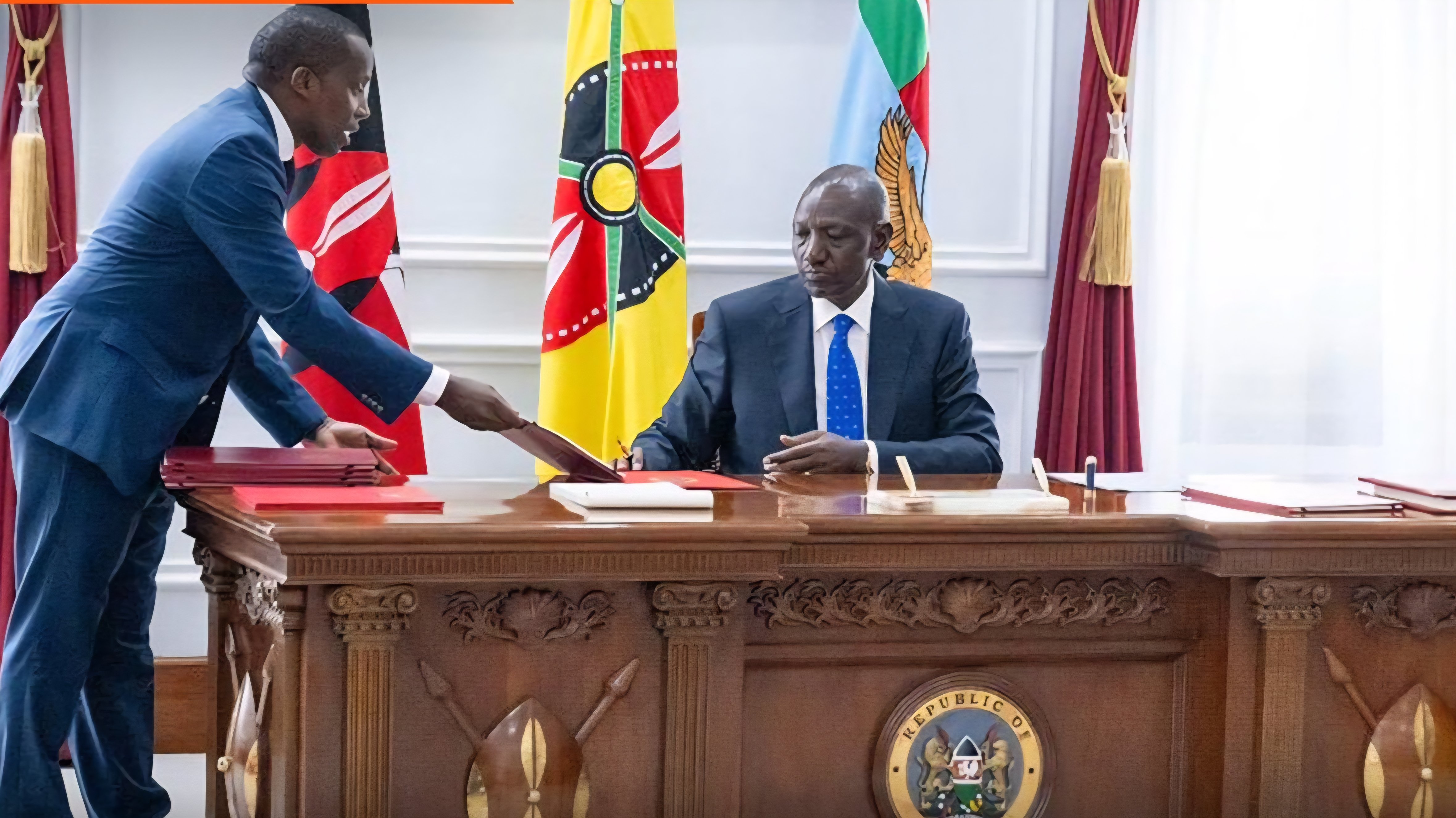 President William Ruto sitting at a formal desk at State House Nairobi while signing a document into law with officials standing nearby.