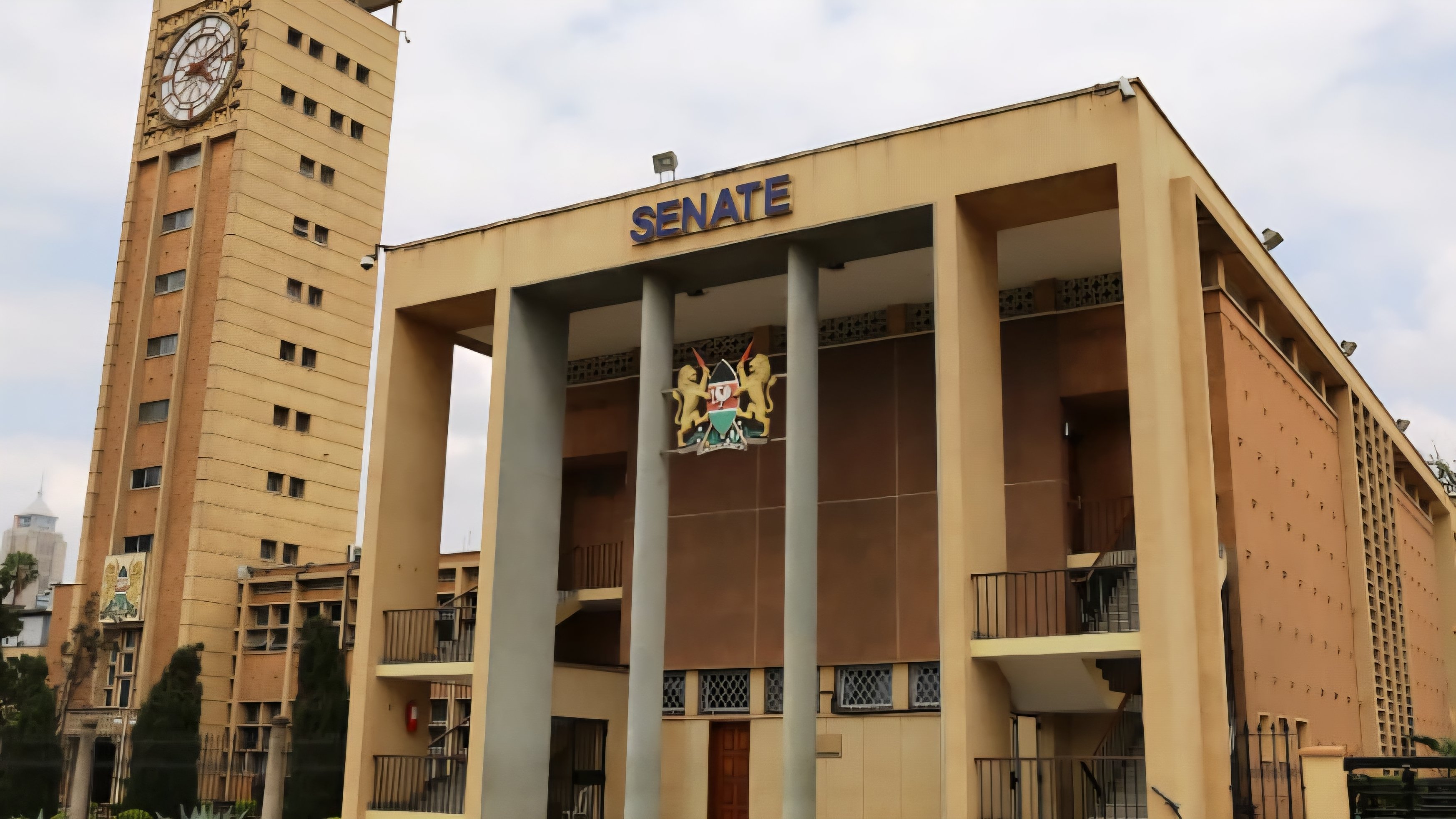 A wide-angle view of the Kenyan Parliament Buildings in Nairobi, featuring the clock tower and the Senate entrance with the national coat of arms displayed prominently above the pillars.