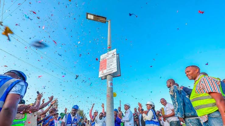 An elevated view of a newly installed solar-powered streetlight pole in a Kenyan town with engineers and officials gathered at the base for an inspection.