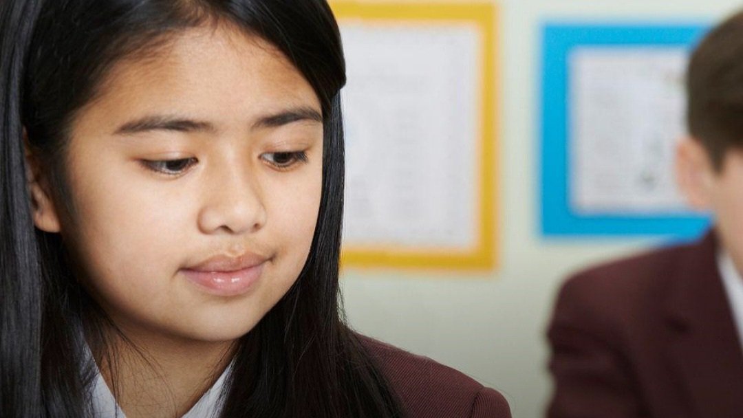A young female secondary school student wearing a brown school uniform jacket sitting in a classroom environment.