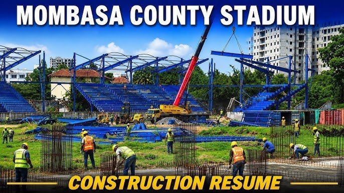 A construction crane lifts blue steel beams at the Mombasa County Stadium site where construction workers are active.