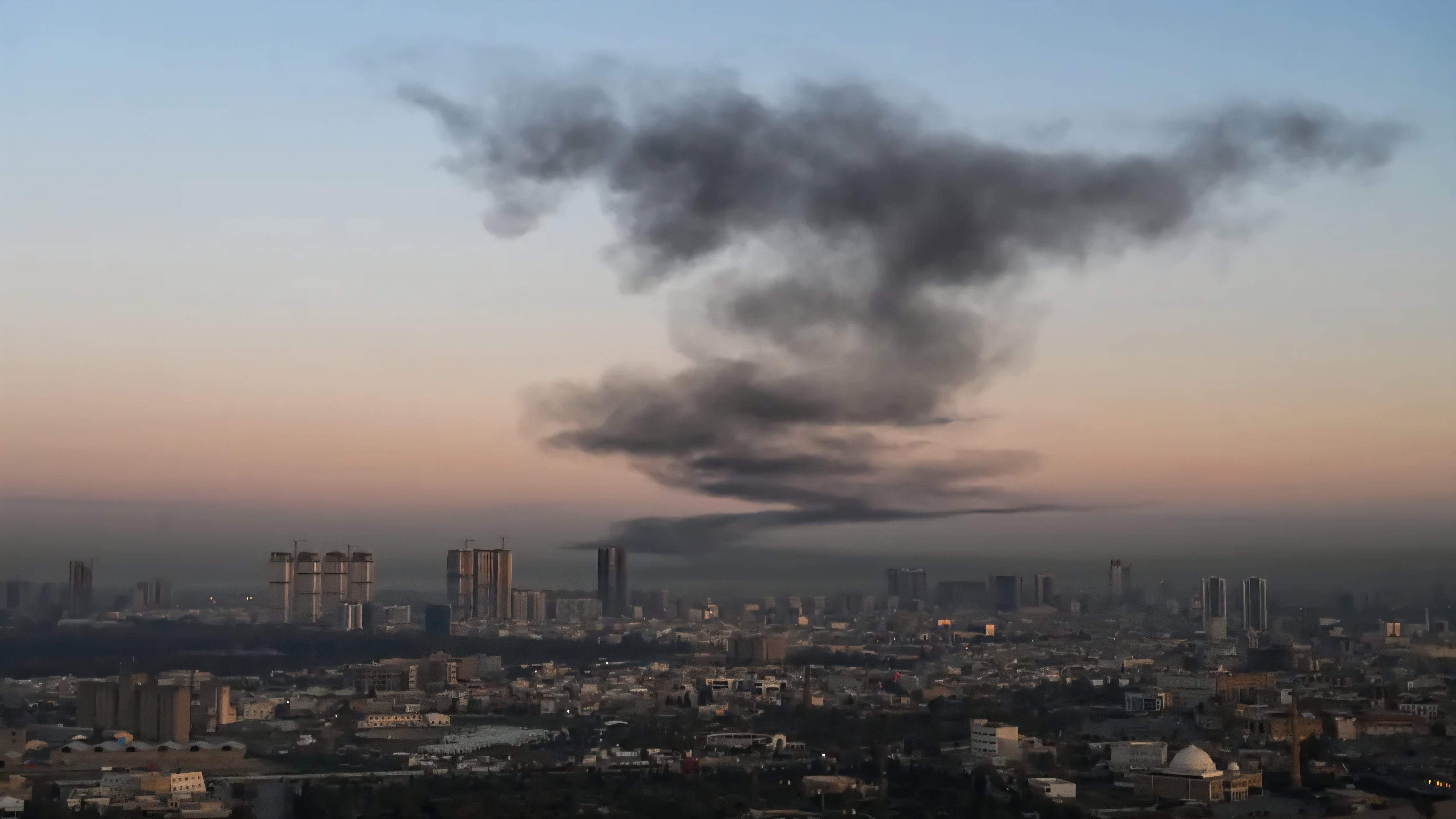 Aerial view of a city skyline with a thick plume of dark smoke rising in the distance under a dusk sky.