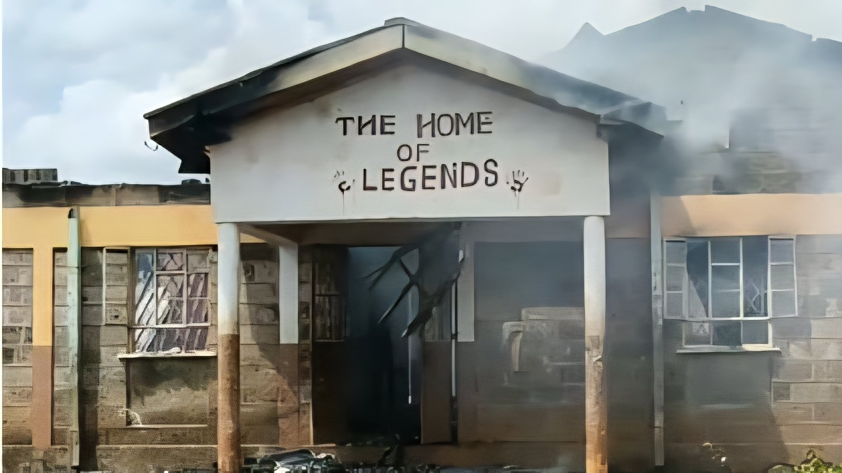 External view of a fire-damaged stone building with smoke billowing from the roof and a sign reading The Home of Legends.