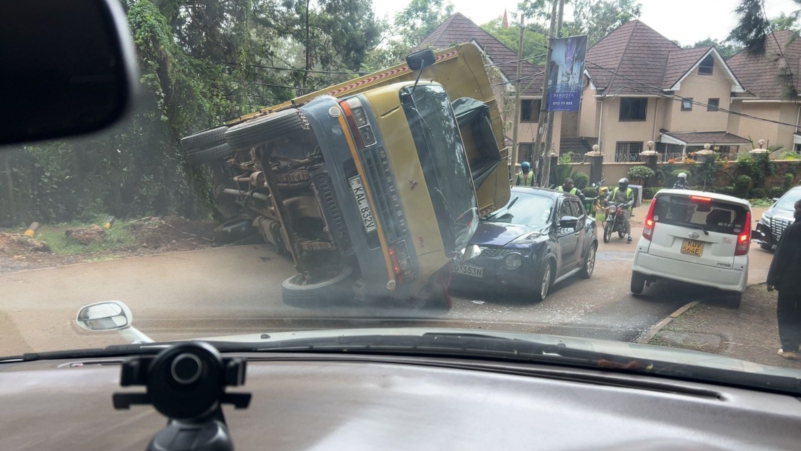 A yellow Mitsubishi tipper truck lying on its side across a paved road, blocking traffic near a residential area with trees in the background.