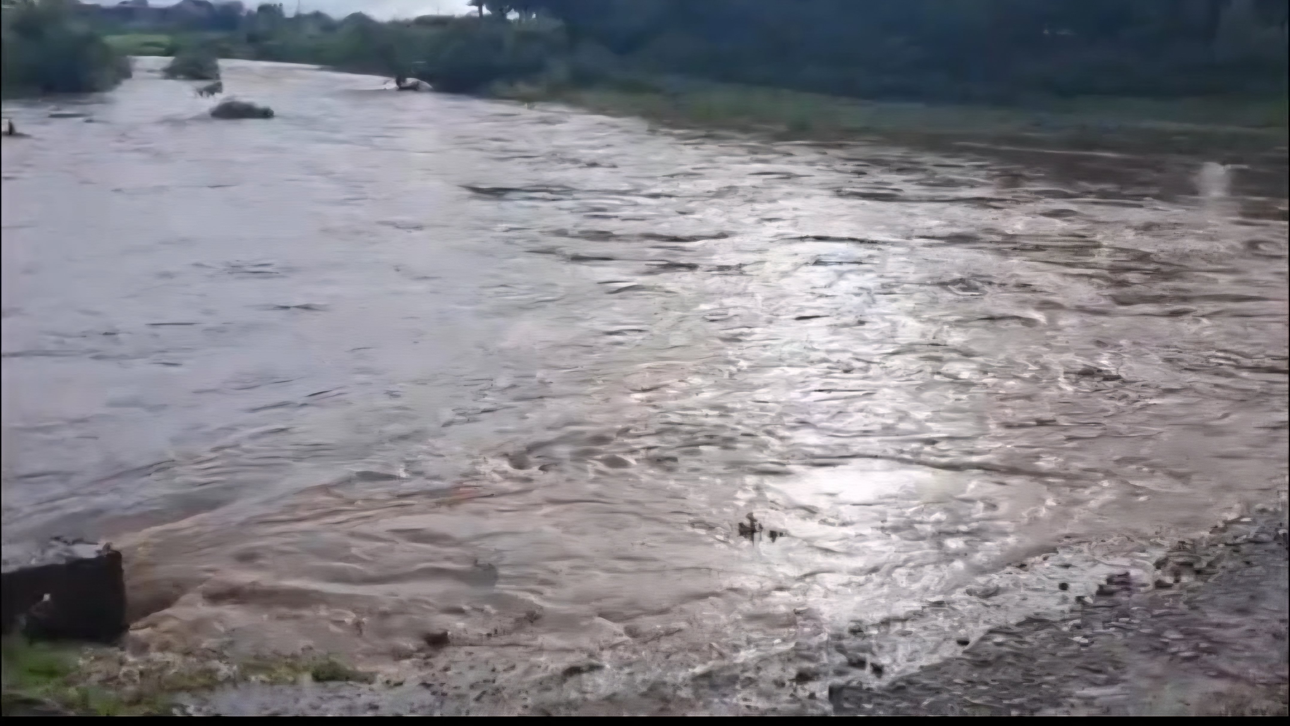 Murky brown floodwaters of River Kanaro flowing rapidly between grassy banks in Kirinyaga County.