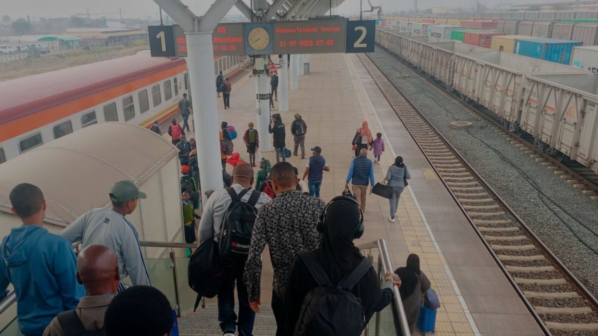 Passengers walking down steps to the platform at a Standard Gauge Railway station in Kenya, with a passenger train on the left and cargo container wagons on the right.