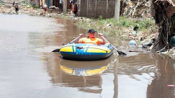 A man sits in a small yellow and blue inflatable boat, paddling through a heavily flooded residential street in Nairobi surrounded by debris.