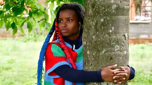 A photo of Kenyan climate activist Truphena Muthoni wearing a colorful crocheted wrap while leaning against a large tree trunk.