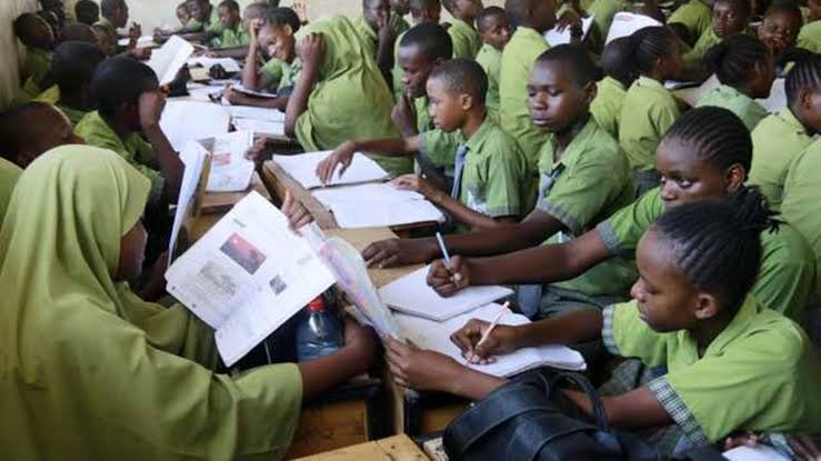 Large group of students in green uniforms seated at wooden desks in a crowded classroom, focused on writing in their notebooks.
