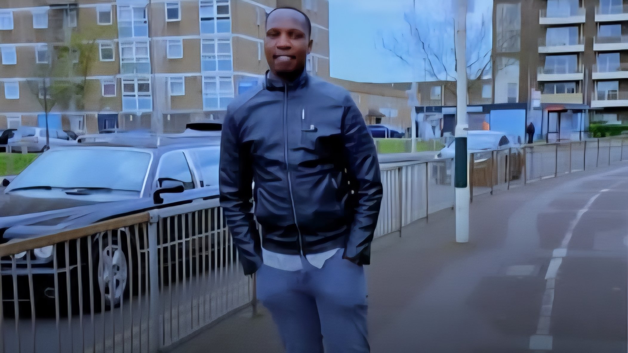 A man in a black leather jacket and grey trousers standing on a London street with residential brick buildings in the background.