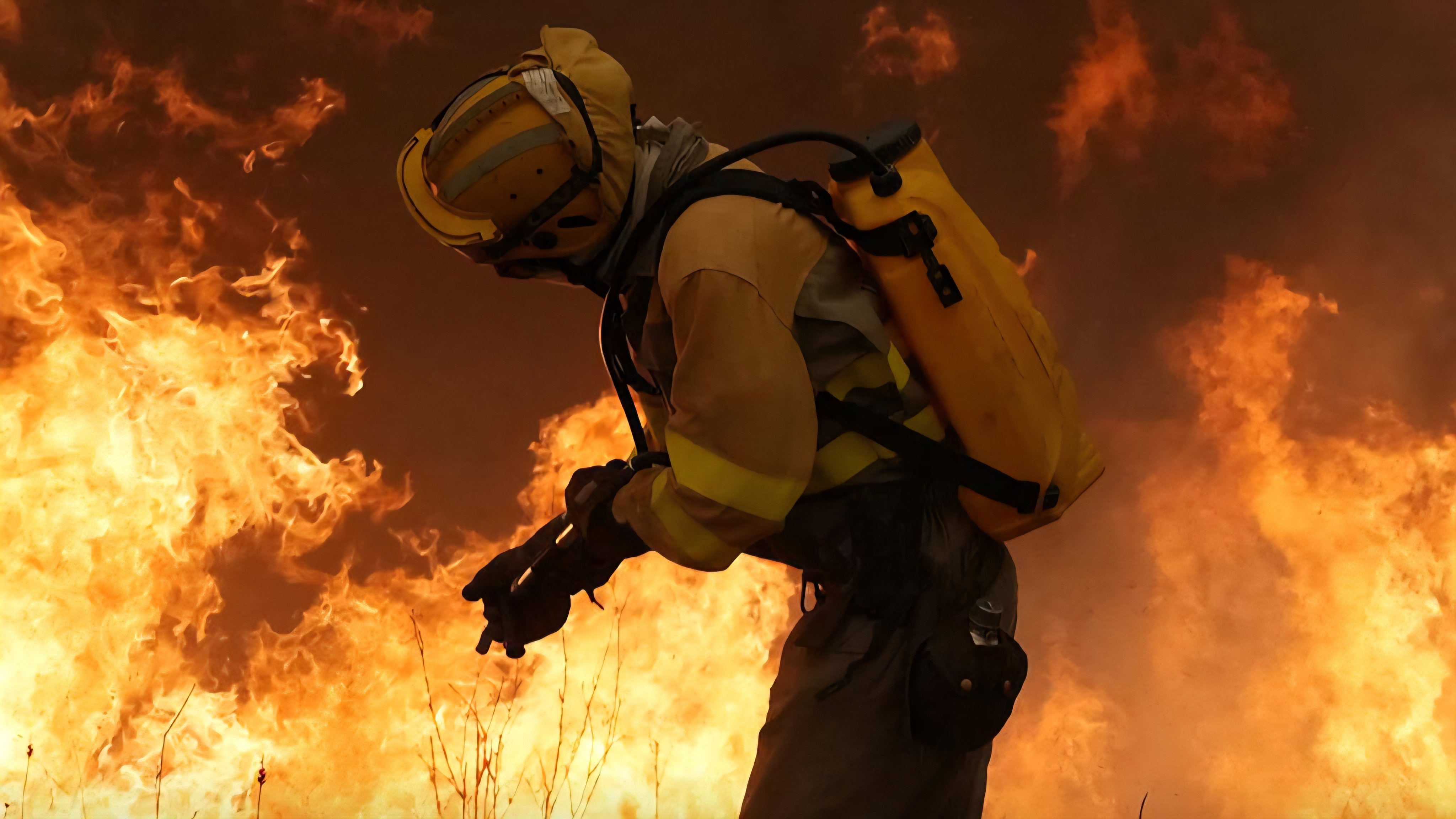 A firefighter carrying a yellow backpack water sprayer walks along a wall of burning fire during a wildfire response operation.