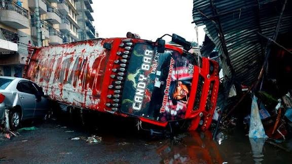 A colorful red and purple matatu bus tipped over onto its side, resting on a silver private car in a flooded street in Nairobi's Grogan area.