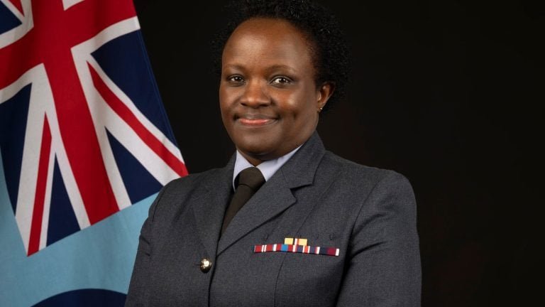 A formal portrait of Group Captain Joan Ochuodho in her Royal Air Force service dress uniform, standing against a dark background with the Union Jack and RAF ensign.