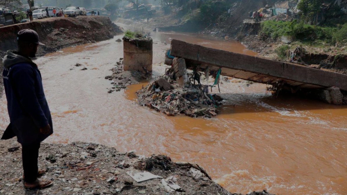 A man standing by a muddy river channel looking at a heavily fractured concrete bridge deck that has collapsed into the water, as depicted in file 288489.png.