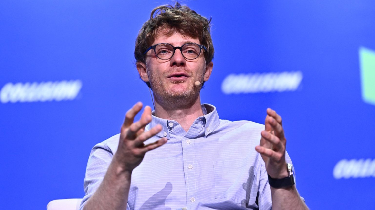 A man with glasses and a light blue button-down shirt speaking on a stage with a blue background.