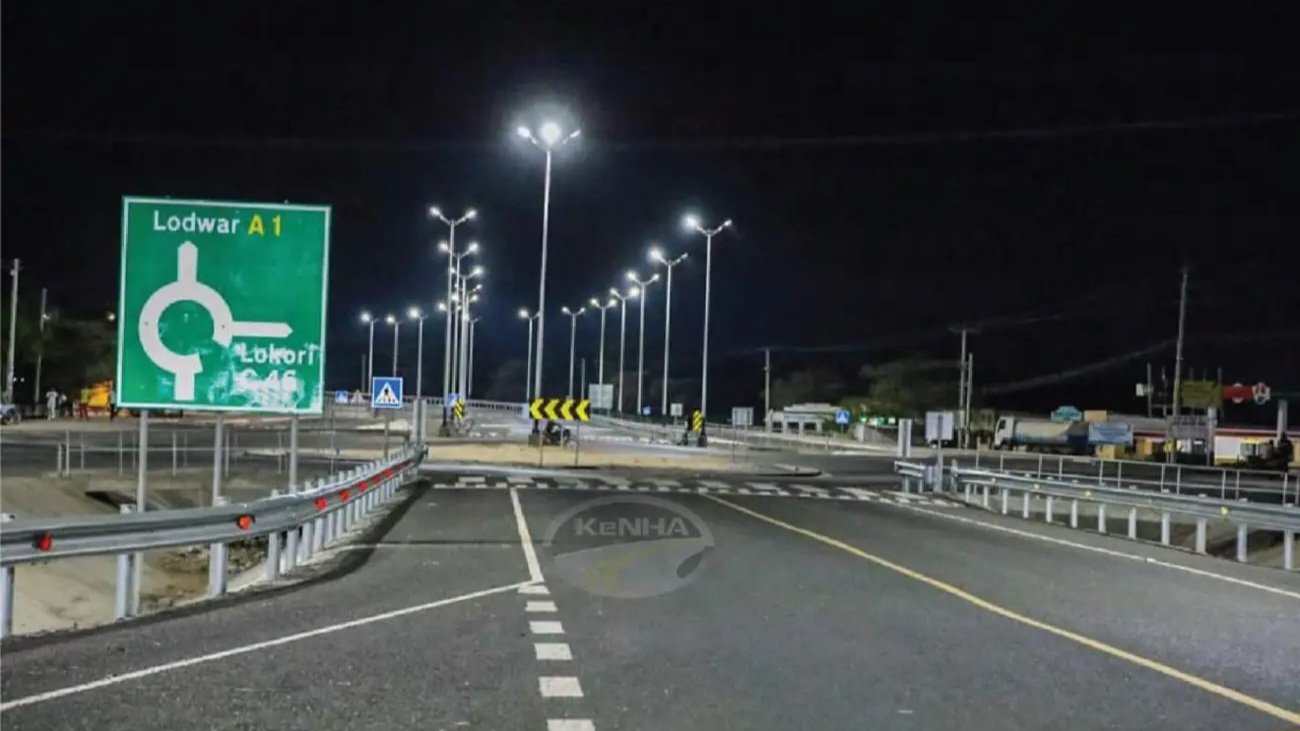 A paved road in Lokichar town with streetlights and local storefronts, showing a motorcycle and pedestrians under a clear sky.