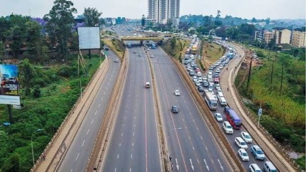 Aerial view of multi-lane highway with vehicles including trucks, vans, and motorcycles traveling in both directions, bordered by grassy median and roadside areas.