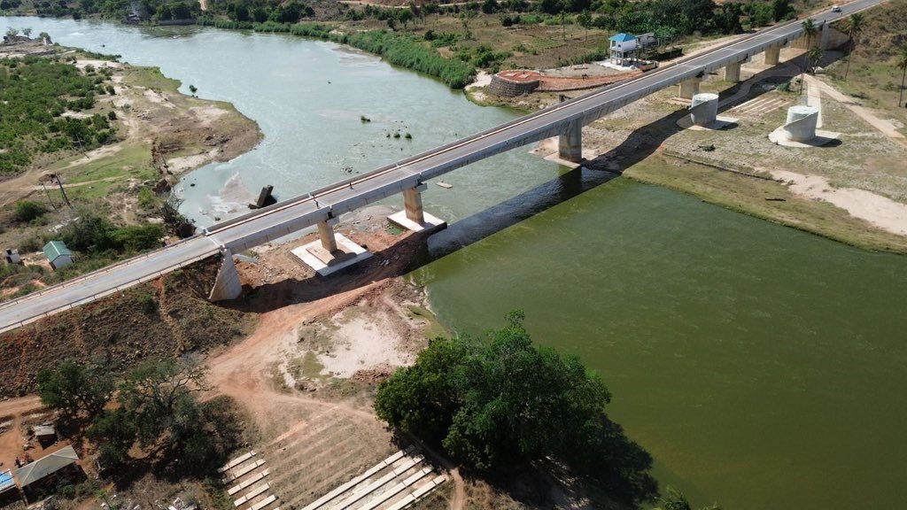 Aerial view of concrete bridge with multiple piers spanning a wide river in rural landscape with green water and surrounding vegetation.