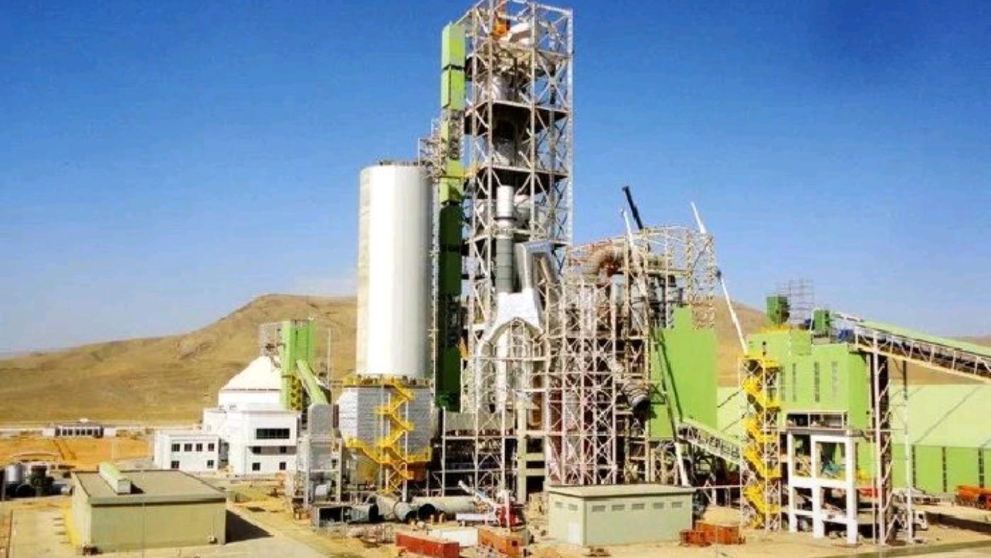A cement factory with storage silos and a large pile of aggregates under a bright sky.