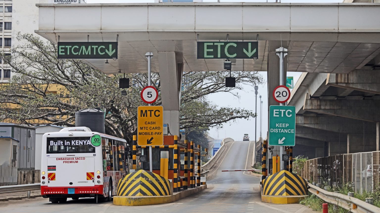 An electric BasiGo bus with "Built in Kenya" branding is seen driving through a toll or security gantry in Nairobi with commercial buildings in the background.