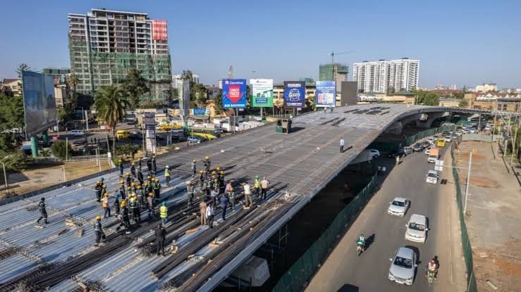 Aerial view and close-up of the Ngong Road-Junction flyover construction site in Nairobi, showing the elevated roadway structure and workers on the bridge deck.