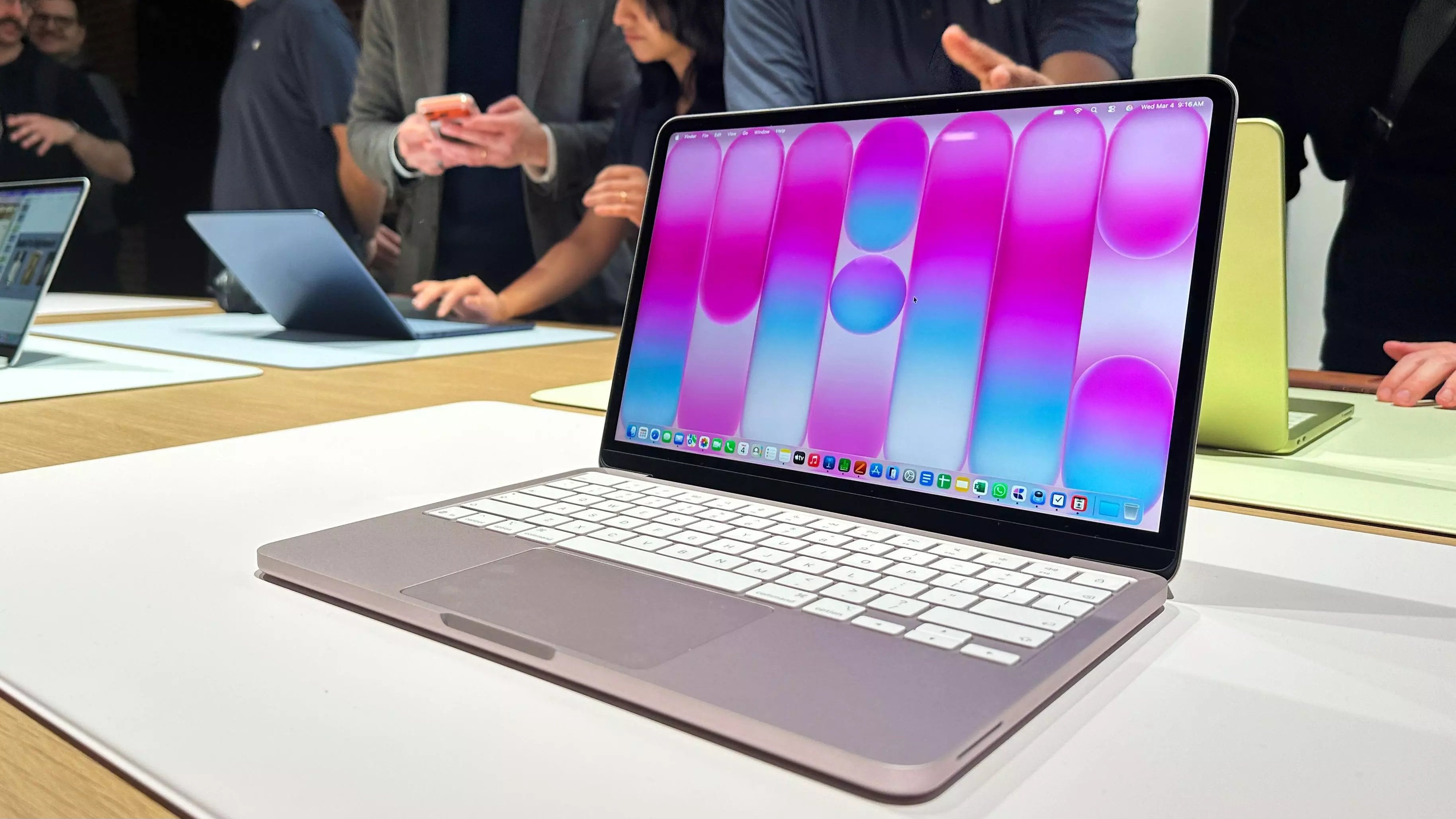 A sleek white-colored Apple laptop sits open on a wooden display table, displaying a vivid red and black high-definition image on its screen, with other laptops visible in the soft-focus background.