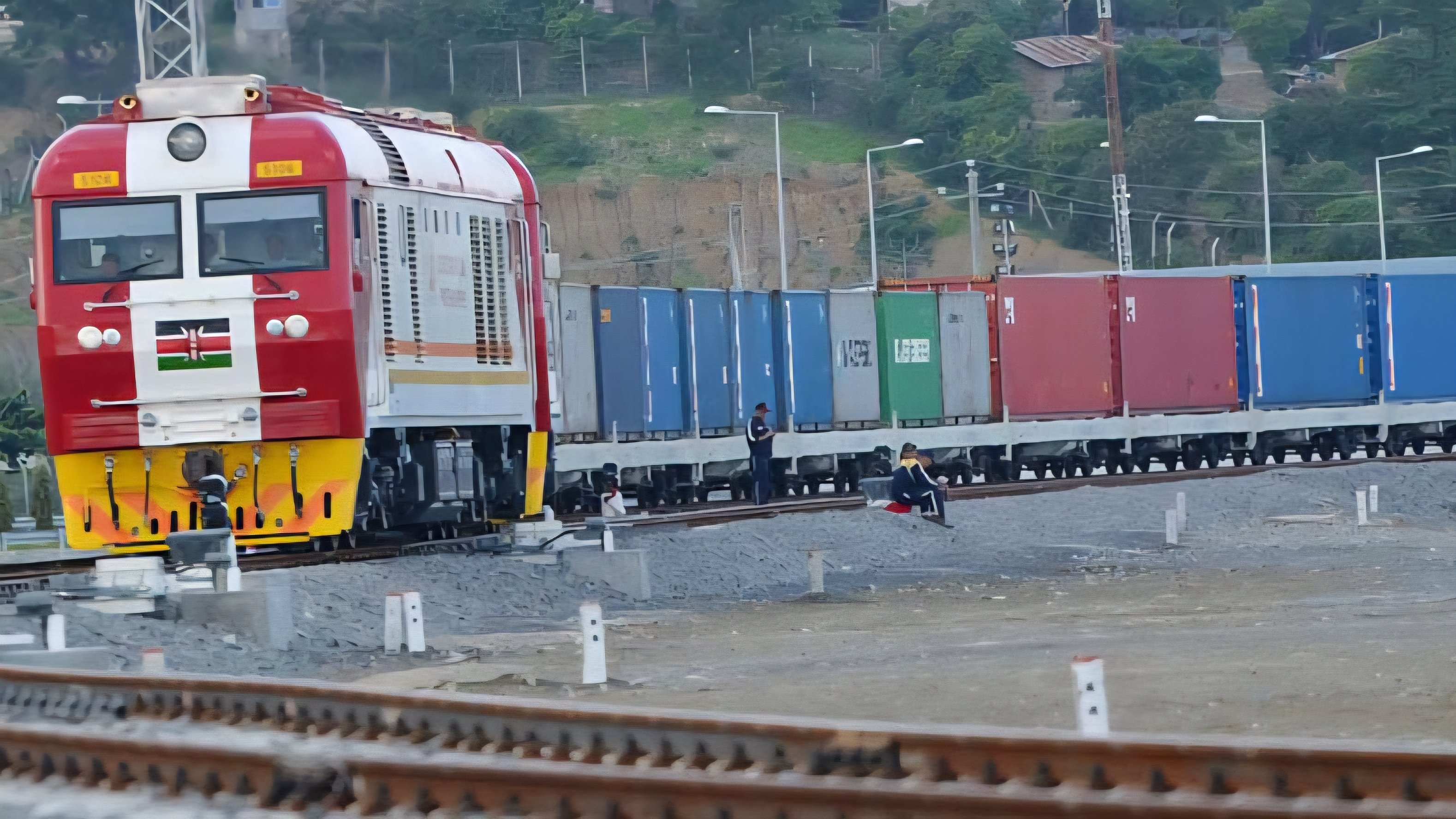 A red and white Kenya Railways cargo locomotive pulling a long line of shipping containers along the Standard Gauge Railway track.
